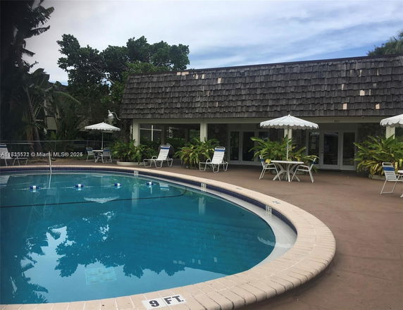 Outdoor swimming pool area with surrounding chairs and tables, adjacent to a building with a shingle roof.