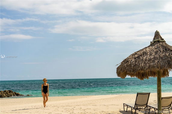 Beach scene with a person walking on sand and a thatched umbrella with lounge chairs.
