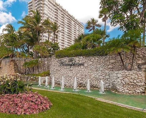 View of a tall residential building with a landscaped area in front featuring a stone wall and water fountains.