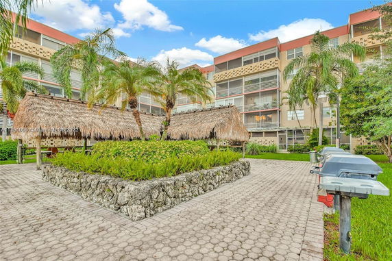 View of an apartment building with outdoor grounds featuring palm trees and thatched pavilions.