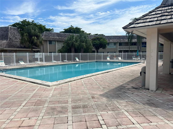 View of an outdoor swimming pool with surrounding paved area and adjacent buildings.