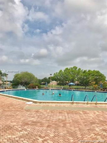 View of a large outdoor swimming pool with people swimming, surrounded by paved area and greenery under a cloudy sky.