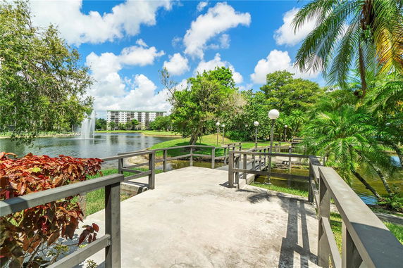 Scenic view of a pond with a fountain surrounded by lush greenery and a multi-story building in the distance.