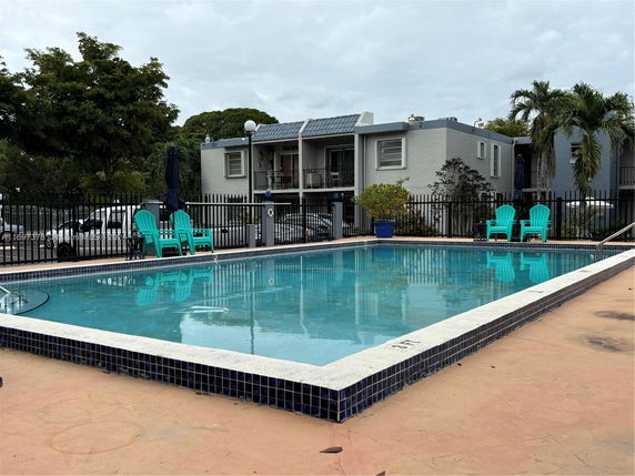 View of a swimming pool with a fence and chairs around it, and a building in the background.