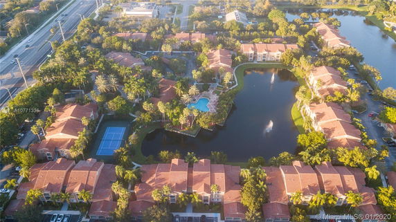 Aerial view of a residential complex with multiple buildings, a central swimming pool, fountains, and a tennis court.