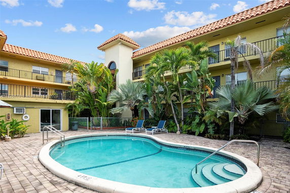 Outdoor swimming pool surrounded by a multi-story building with balconies and tropical plants.