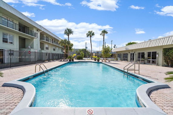 Outdoor swimming pool with surrounding buildings and palm trees.