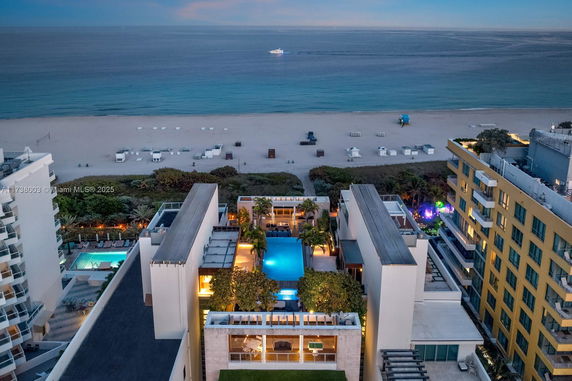 Aerial view of a coastal area with buildings surrounding a central swimming pool, and a beach in the background.