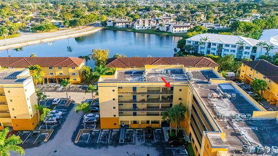 Aerial view of a multi-story apartment building with a flat roof, surrounded by parking spaces and nearby water body.