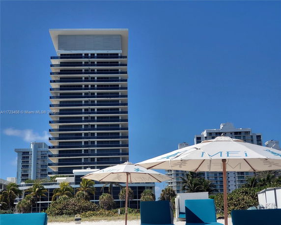 A high-rise building with multiple balconies against a clear blue sky, surrounded by other structures.
