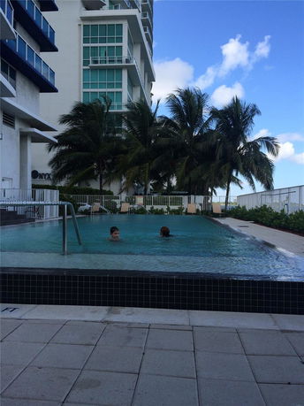 View of a swimming pool next to a multi-story building with large windows and palm trees nearby.