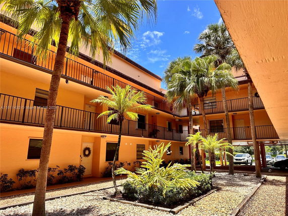 Outdoor view of an apartment complex with several floors and balconies, surrounded by palm trees and a paved courtyard.