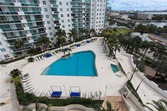 View of a high-rise condominium building with a large outdoor swimming pool area surrounded by palm trees.
