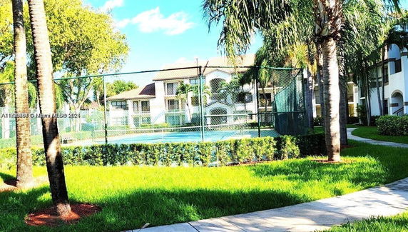 View of an outdoor tennis court surrounded by palm trees and residential buildings.