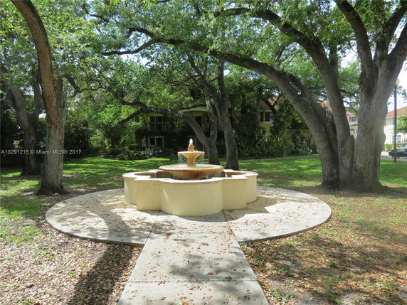 View of a yellow house with multiple windows surrounded by large trees and a water fountain in front.