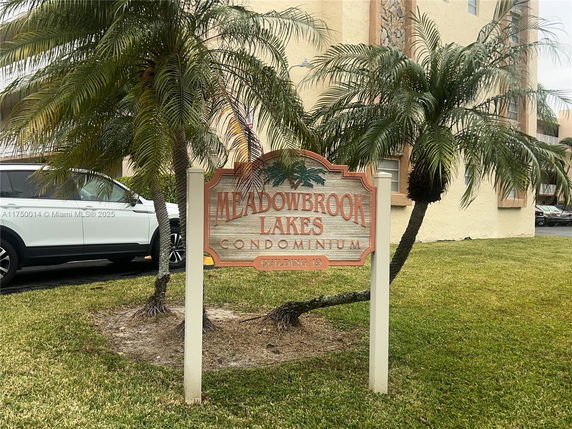 Sign for Meadowbrook Lakes Condominium with a building and palm trees in the background.