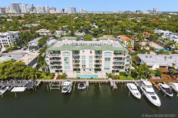 Aerial view of a multi-story building with balconies and a pool, located next to a waterfront with several boats docked.