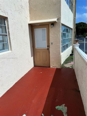 Exterior view of a beige building with red flooring and a brown door.