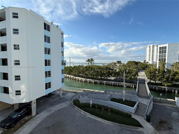View of mid-rise buildings near a waterfront with a walkway and trees in the background.