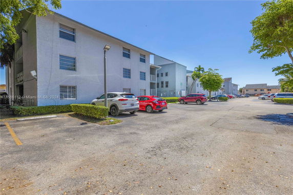 Outdoor parking area with several cars and a multi-story building in the background.
