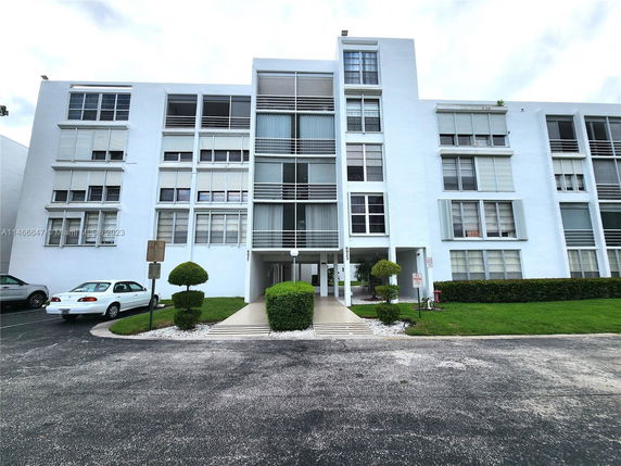Front view of a multi-story residential building with balconies and a parking area in front.