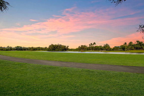 Scenic view of a grassy area with a path, palm trees, and a lake under a sunset sky.