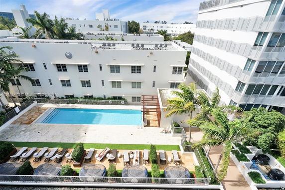 View of a swimming pool area beside a multi-story building with lounge chairs and palm trees.