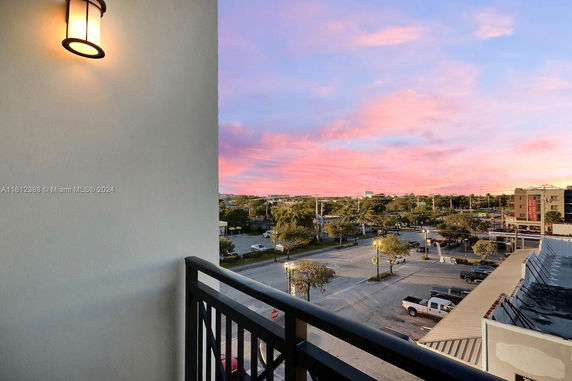 Panoramic view from a building balcony overlooking a parking area and trees against a sunset sky.