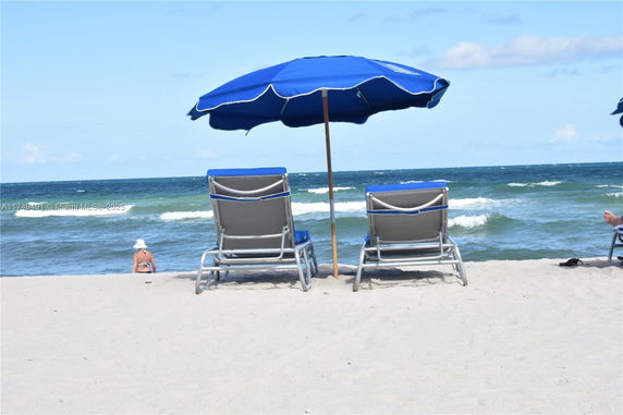 Beach scene with two lounge chairs under a blue umbrella, facing the ocean.