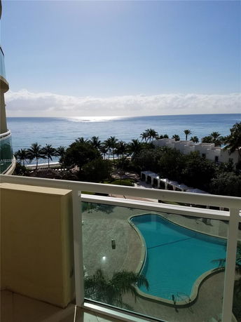 View from a balcony overlooking a swimming pool and the ocean with palm trees.