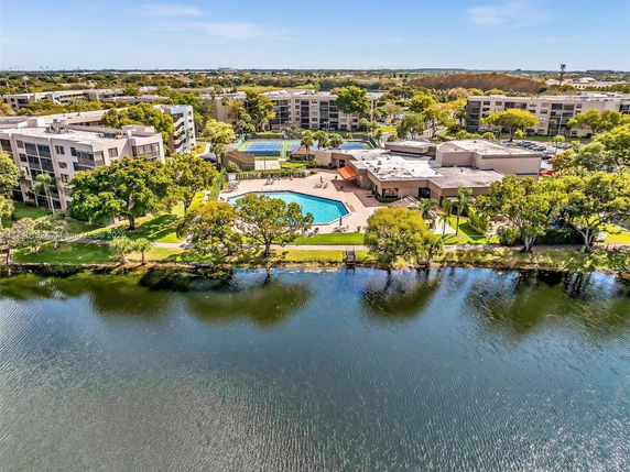 Aerial view of buildings with a swimming pool and tennis court surrounded by trees and a waterbody.