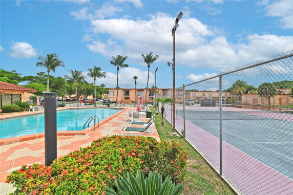 Outdoor swimming pool with adjacent tennis court and buildings in the background.
