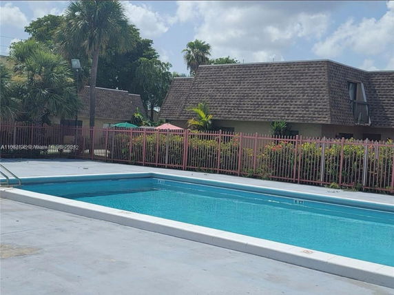 View of an outdoor swimming pool with a residential building featuring a shingled roof and small balconies in the background.