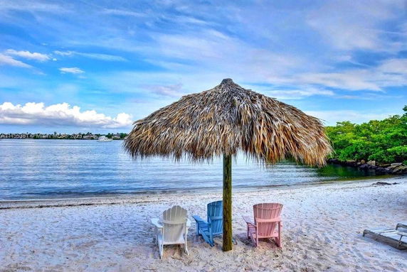A beach scene with a thatched umbrella and three colorful Adirondack chairs on the sandy shore.