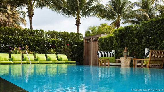 Outdoor area with a pool, surrounded by palm trees and greenery, featuring green lounge chairs and a small seating area.