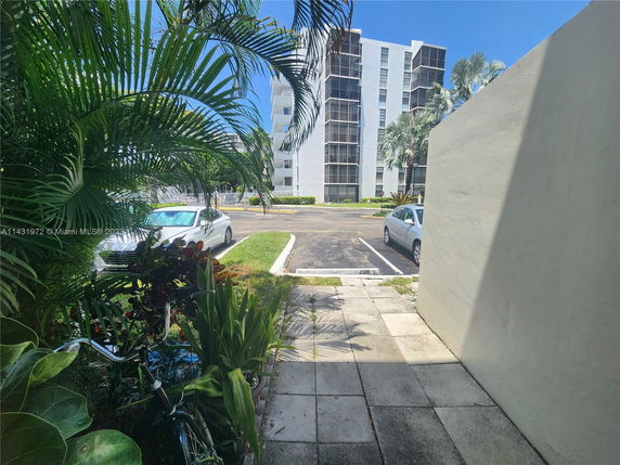 Outdoor parking area near a multi-story building with parked cars and surrounding plants.