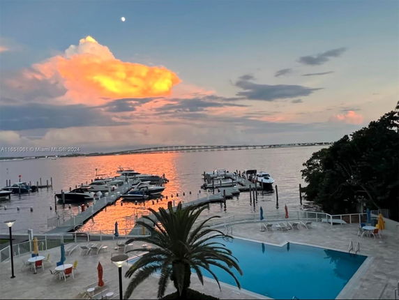 View over a marina with several docked boats and a swimming pool area in the foreground, under a colorful sunset sky.