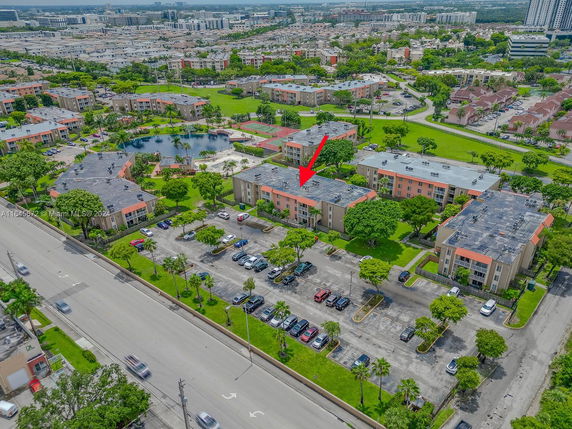 Aerial view of a residential complex with multiple buildings and a parking lot, surrounded by greenery and roads.