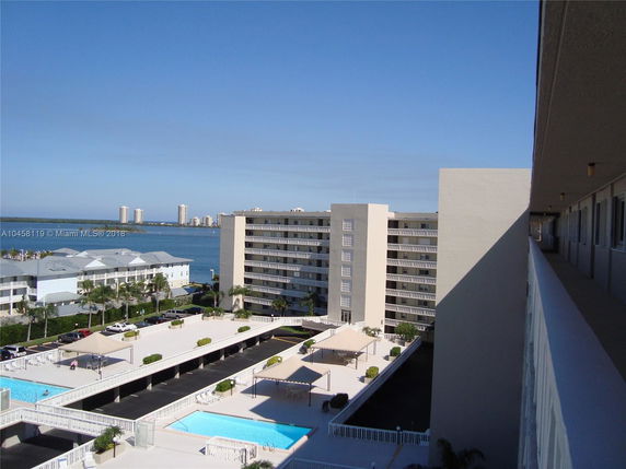 View from a building's balcony overlooking a pool area and other residential buildings with a body of water and distant cityscape in the background.