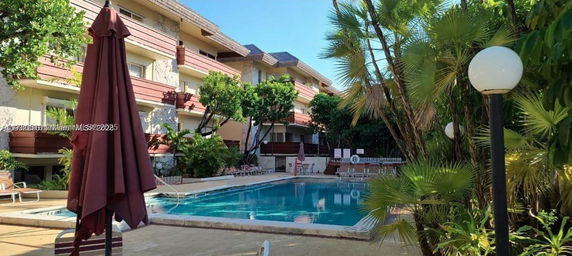 Outdoor view of a swimming pool area with adjacent multi-story buildings and palm trees.