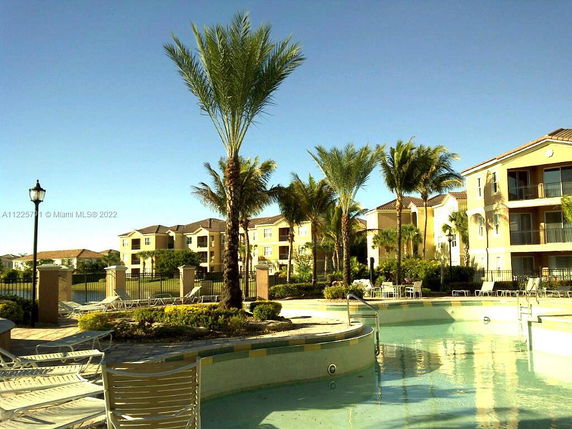 Outdoor area with a swimming pool surrounded by palm trees and multi-story buildings in the background.