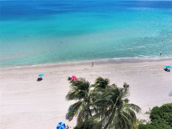 View of a sandy beach with turquoise ocean waters, palm trees, and scattered beach umbrellas.
