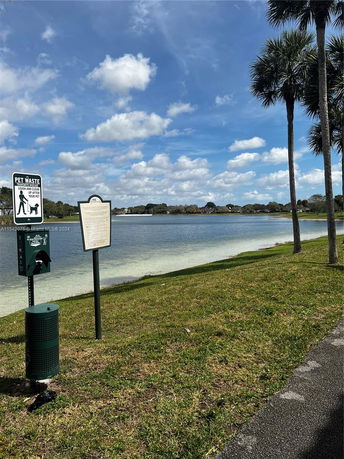 A view of a lake with a grassy area and palm trees, along with a pet waste station and sign.