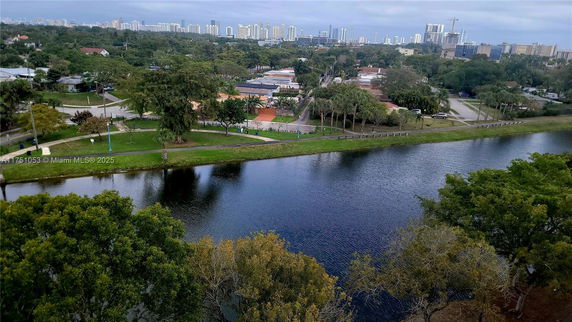 Aerial view of a water body surrounded by greenery and residential buildings with a city skyline in the background.