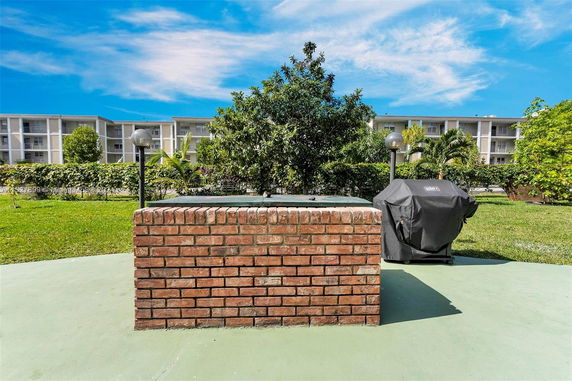 Outdoor area with a brick structure and a covered grill, surrounded by greenery and apartments in the background.