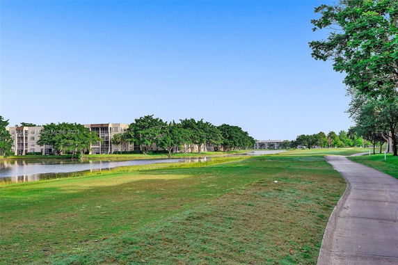 Pathway alongside a grassy area with a view of buildings and trees across a water body.