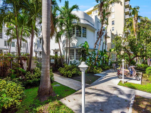Front view of a white multi-story building with art deco architectural details surrounded by tropical plants and trees.