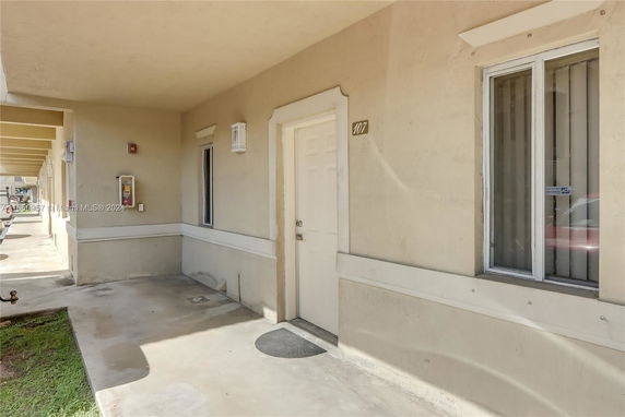 Exterior view of a building entrance with a beige facade, a door marked '107', and a window with vertical blinds.
