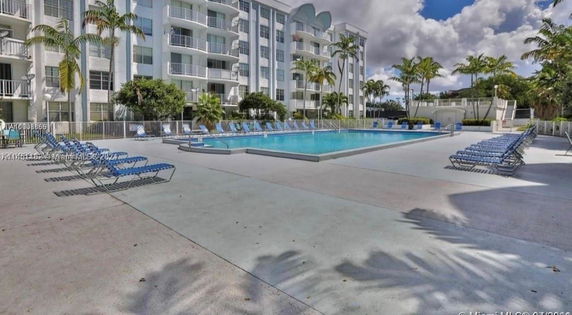 Outdoor swimming pool area with seating and a multi-story building in the background.