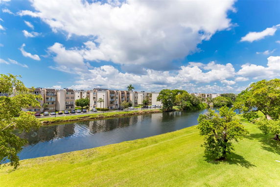 View of a series of four-story buildings alongside a river with greenery under a blue sky.